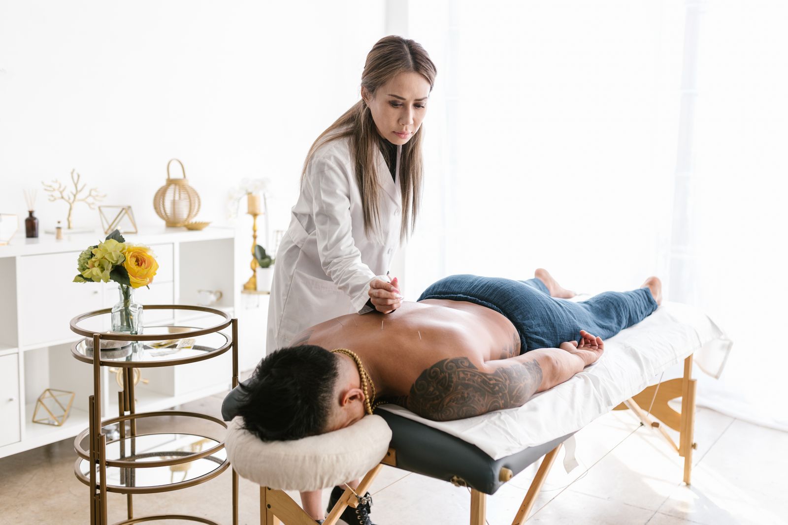An acupuncturist in a white coat treating a patient lying on a table in a bright, uncluttered treatment room