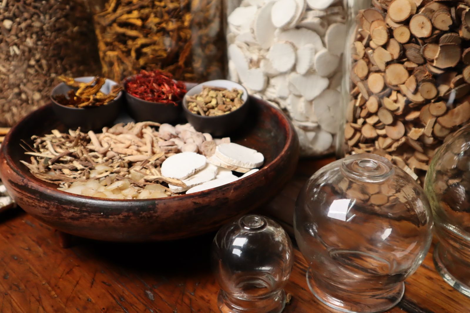 A wooden bowl of dried Chinese herbs and roots beside glass cupping jars