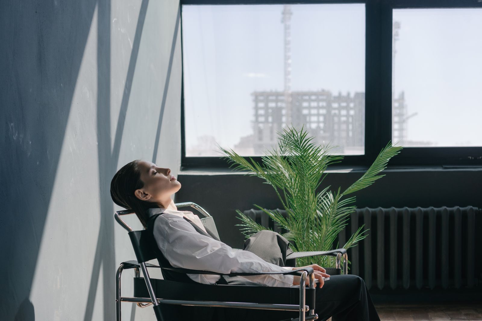 A woman resting with her head back and her eyes closed in a chair by a sunny window, beside a leafy plant