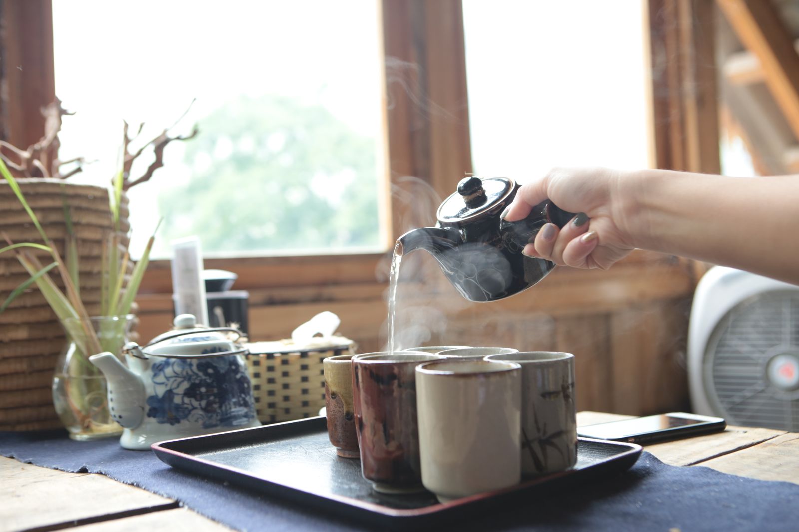 A hand pouring hot tea into small ceramic cups on a tray beside a sunny window